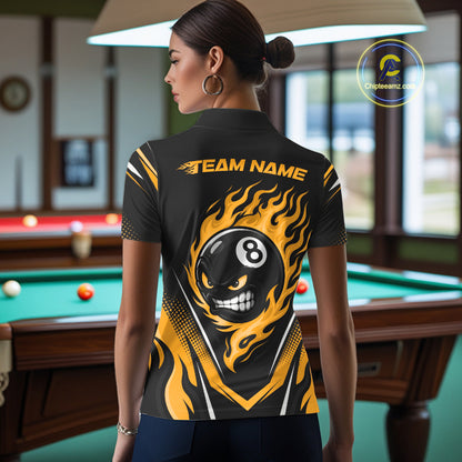 A woman stands in a billiards room wearing a black short-sleeve polo shirt with a bold yellow and black flame-and-billiards graphic on the back. The shirt features a large black 8-ball skull design with yellow flames