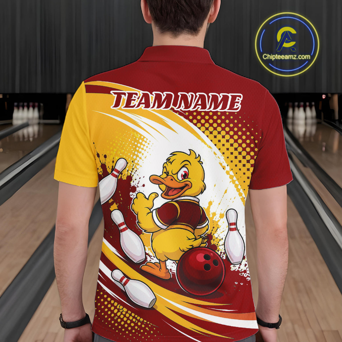 A person viewed from behind wearing a short-sleeve polo shirt in a bowling alley. The shirt has a red and yellow color-blocked design with a large cartoon duck graphic on the back, bowling pins, bowling balls, and the text 'TEAM NAME'.