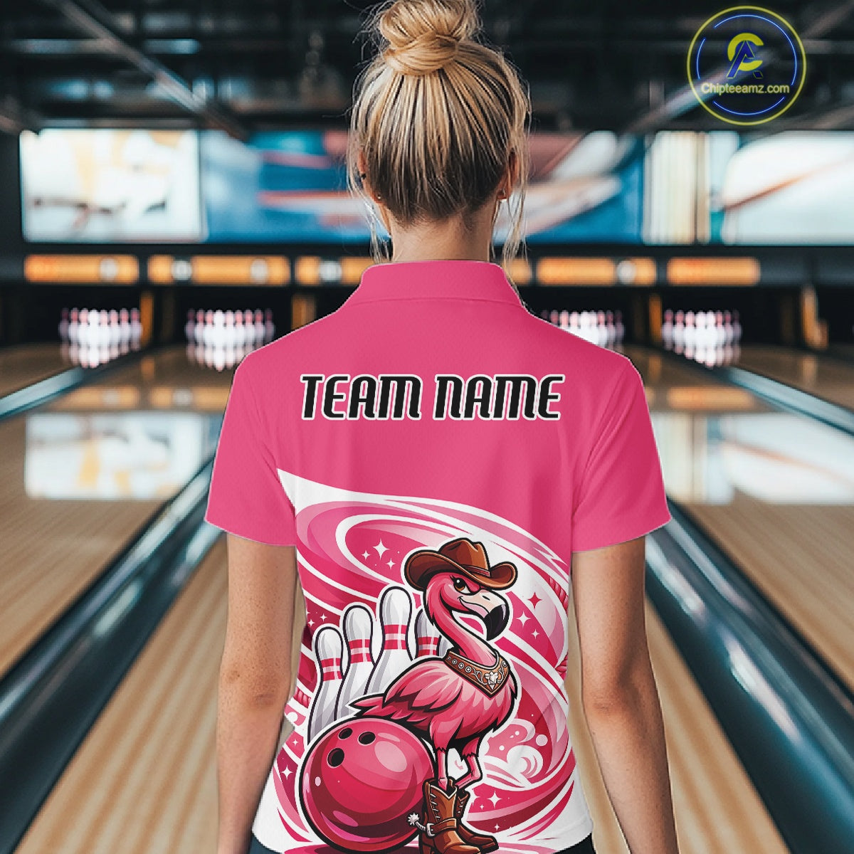 Back view of a female model in a bowling center wearing a pink quarter-zip shirt featuring a cowboy flamingo leaning on a bowling ball.