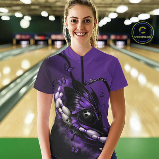 Female model at a bowling lane wearing a purple and black quarter-zip bowling shirt with butterfly artwork embracing a bowling ball and pins.