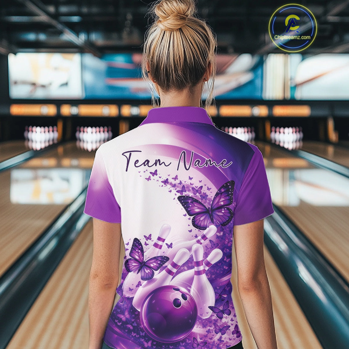 Female model wearing a purple bowling polo with butterfly artwork and bowling graphics, back view at the bowling lane