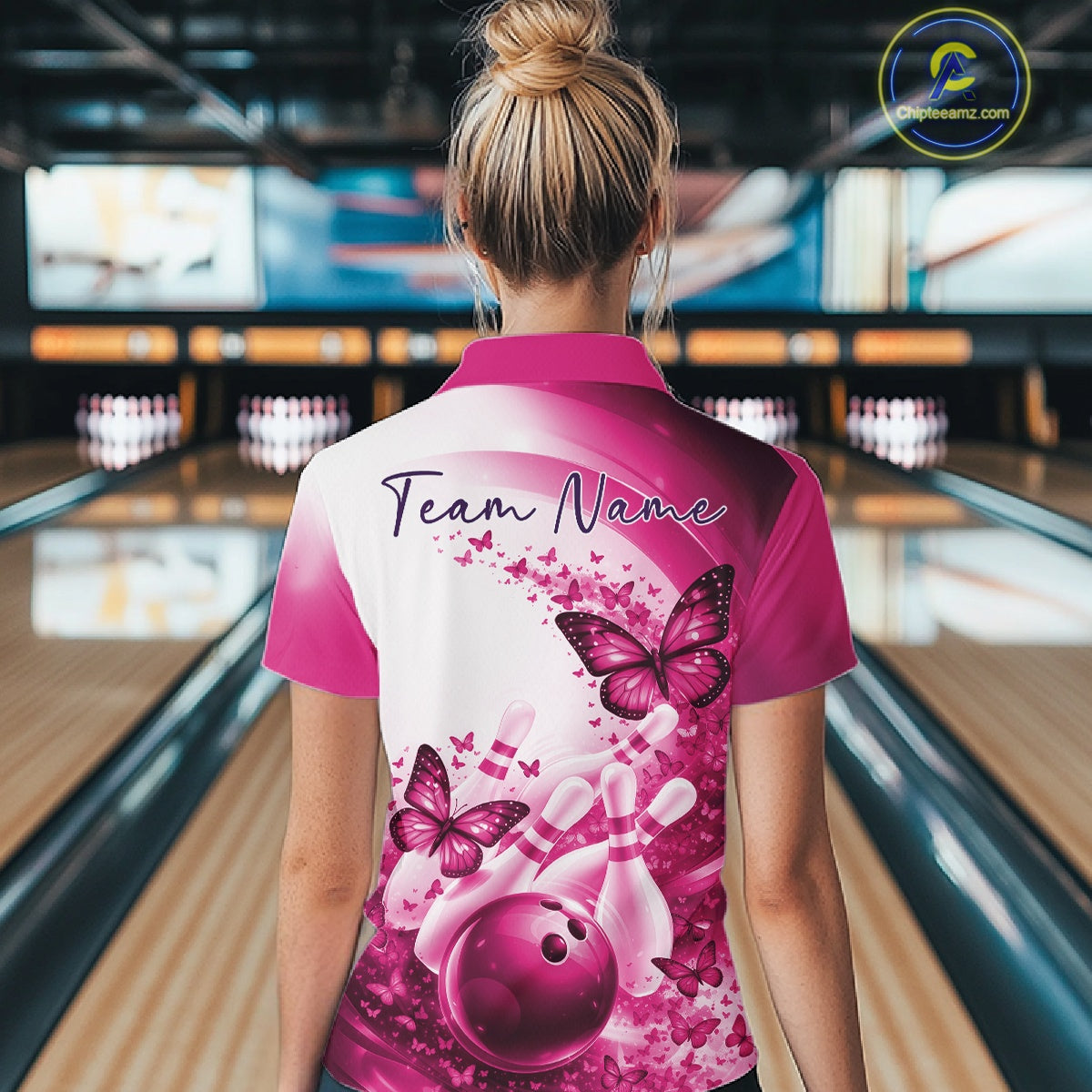 Back view of a female model wearing a pink bowling quarter zip with butterfly artwork and bowling graphics at the bowling lane
