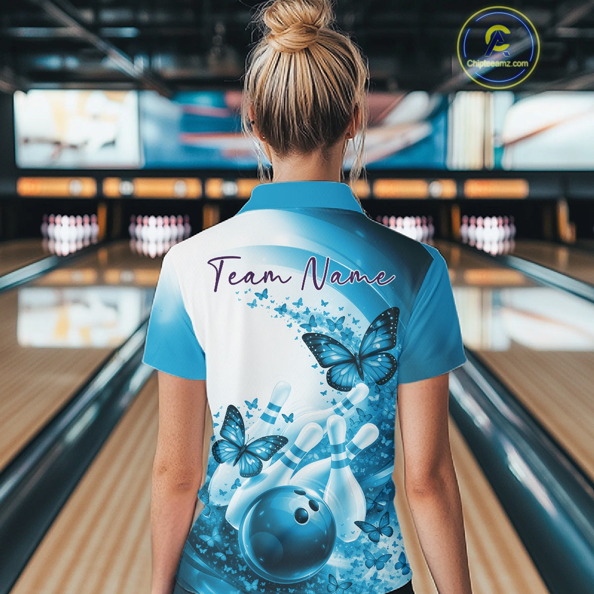 Female model in a blue bowling polo with butterfly artwork and bowling graphics, back view inside a bowling alley