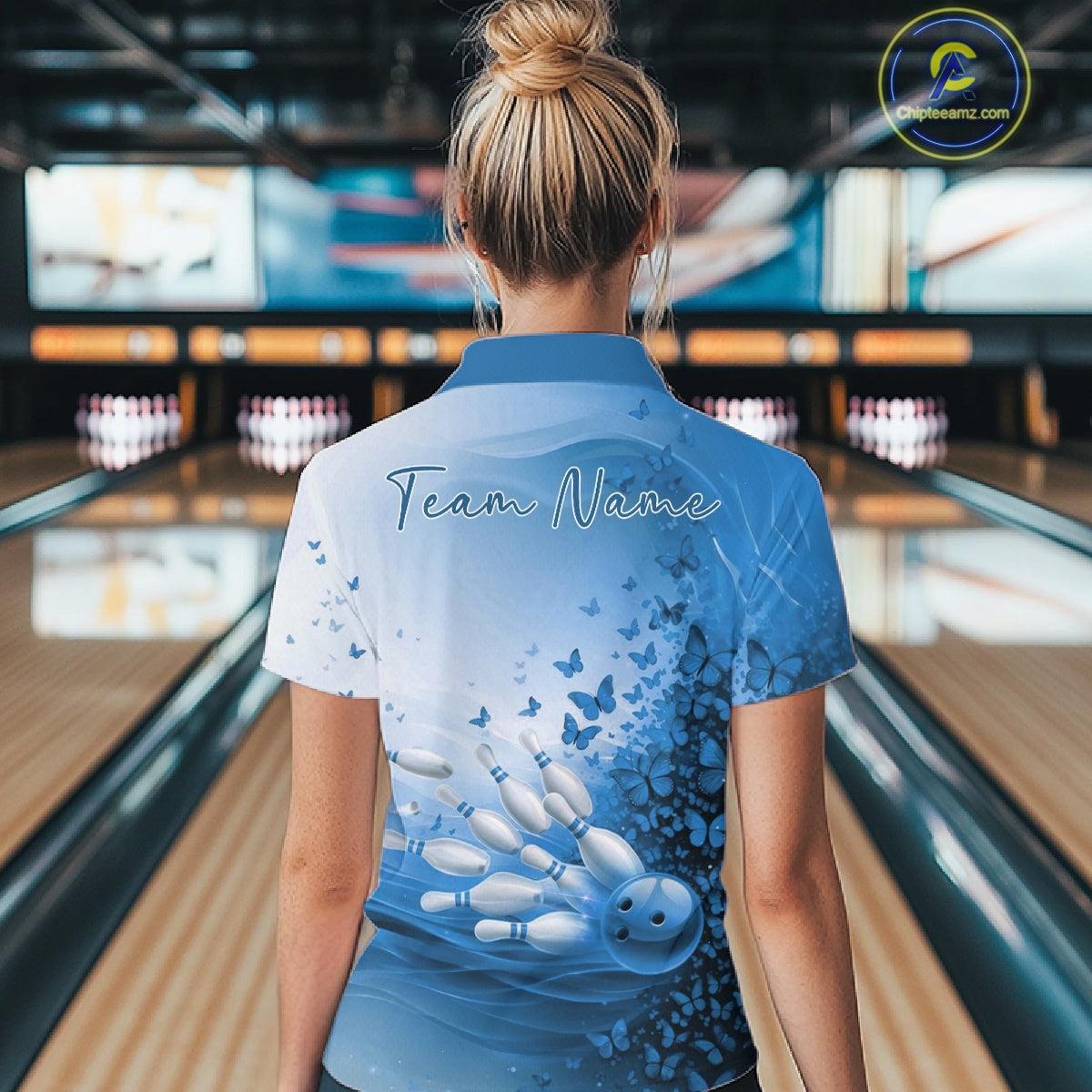 Back view of a white and blue women s bowling polo shirt showcasing butterfly artwork and bowling-themed graphics inside a bowling center.