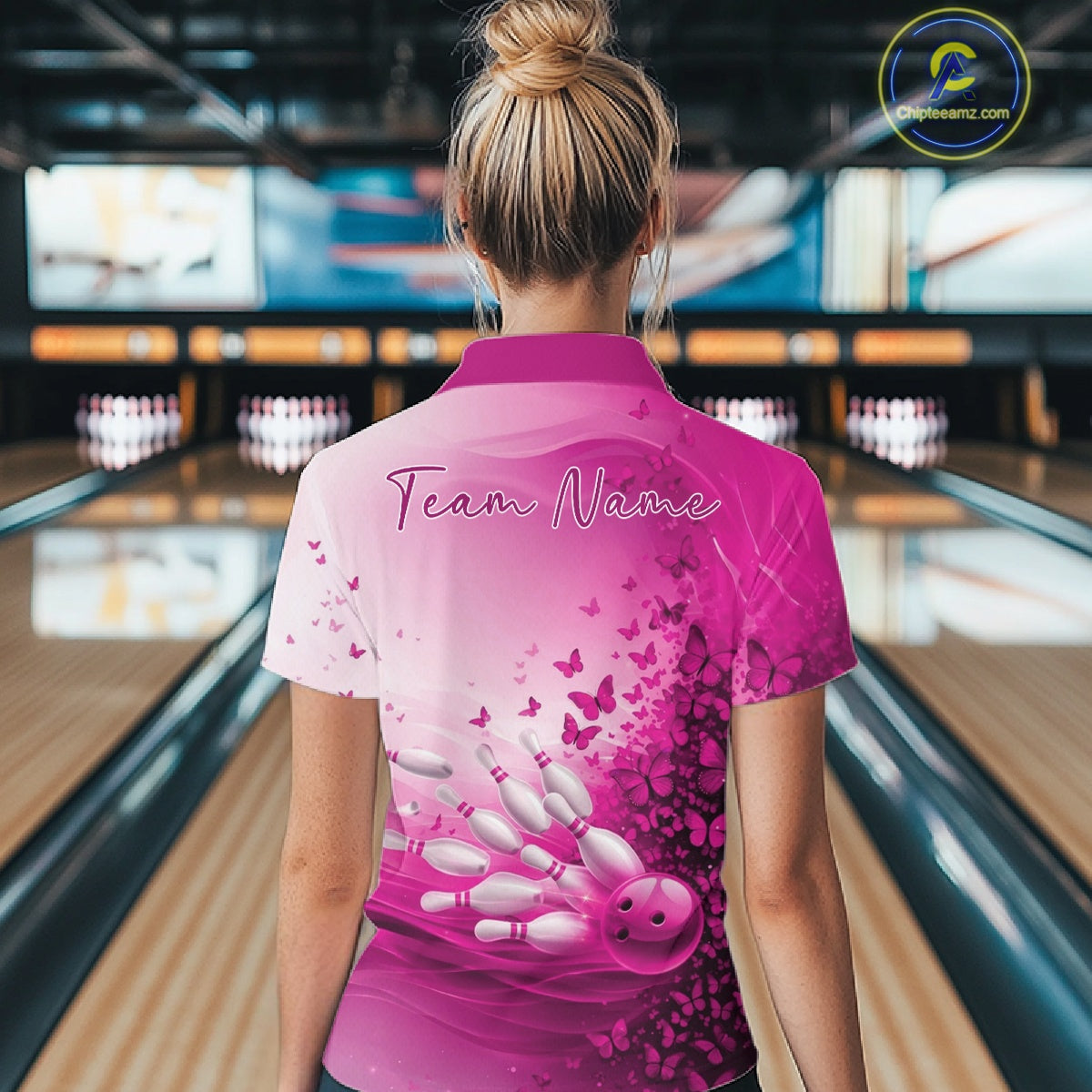 Back view of a women s white and pink bowling polo shirt displaying butterfly accents and bowling graphics inside a bowling alley.