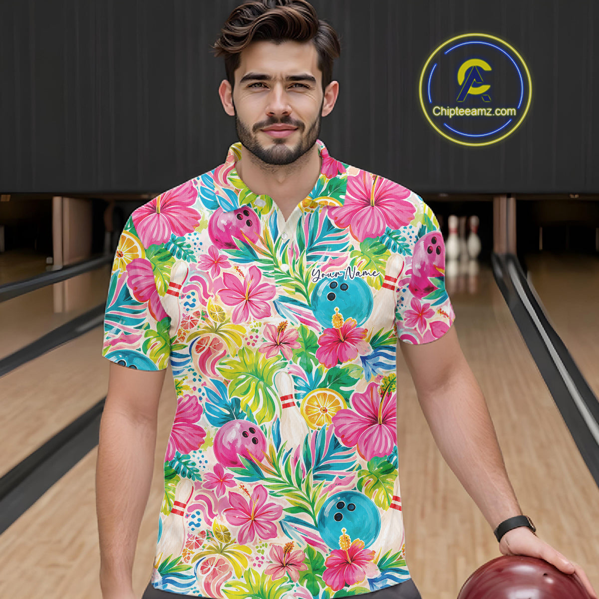 A man wearing a short-sleeve bowling polo shirt with a vibrant tropical floral and hibiscus print, standing in a bowling alley. The shirt features pink flowers, green leaves, blue bowling balls, and palm trees on a cream background.