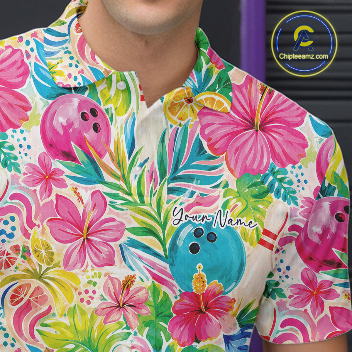 A close-up of a man wearing a short-sleeve collared polo shirt with a vibrant tropical print featuring pink hibiscus flowers, green palm leaves, bowling pins, and citrus slices on a cream background. 