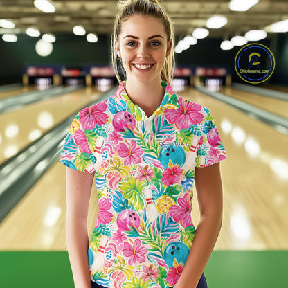 A woman stands in a bowling alley wearing a short-sleeve bowling polo shirt with a bright tropical floral print. The shirt features pink hibiscus flowers, green palm leaves, blue bowling balls, and bowling pins on a white background.