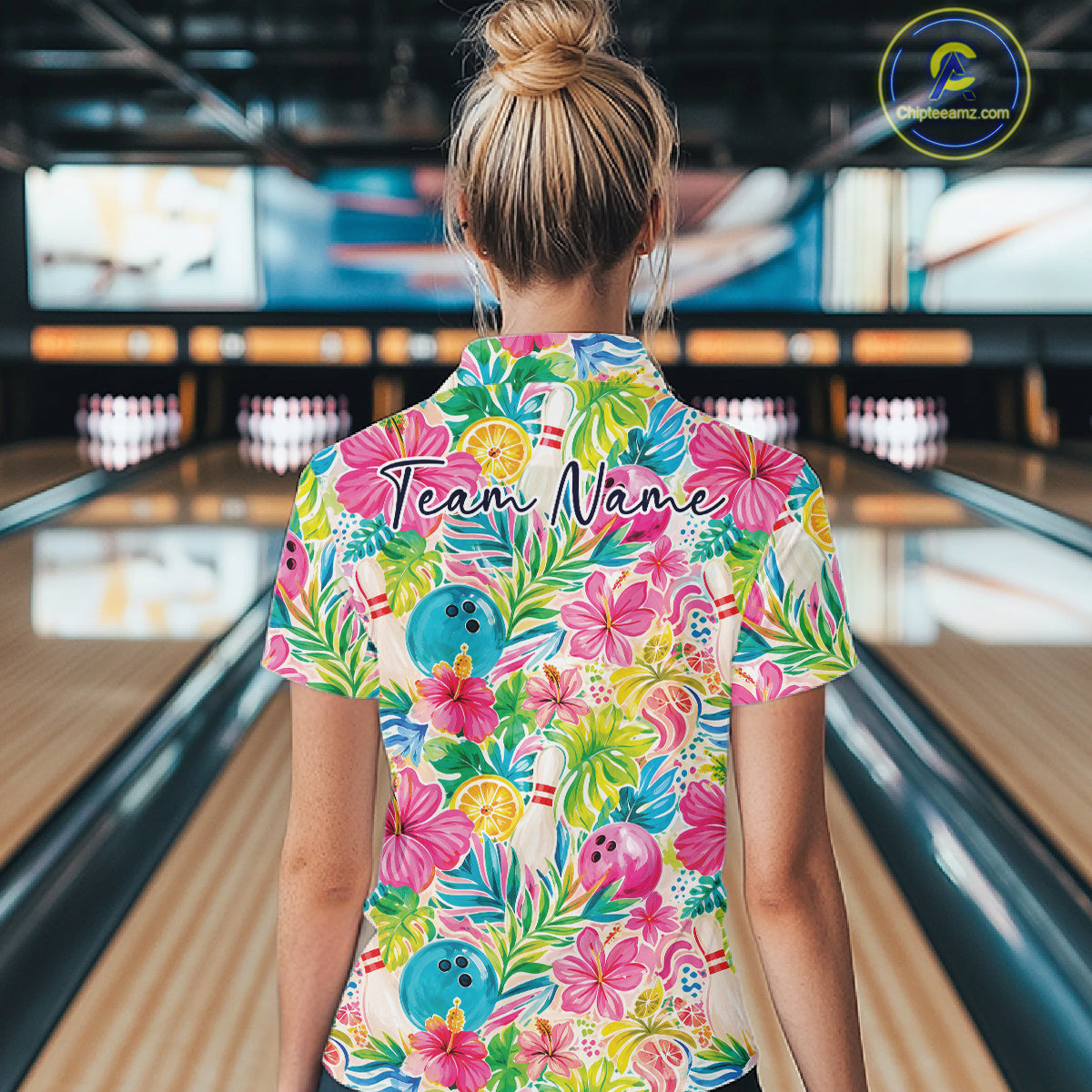 A woman viewed from behind in a bowling alley, wearing a short-sleeve collared polo shirt with a colorful tropical floral print featuring pink hibiscus flowers, green leaves, blue bowling balls, and orange citrus slices. The shirt has a custom name printed across the upper back reading 'Team Name'.