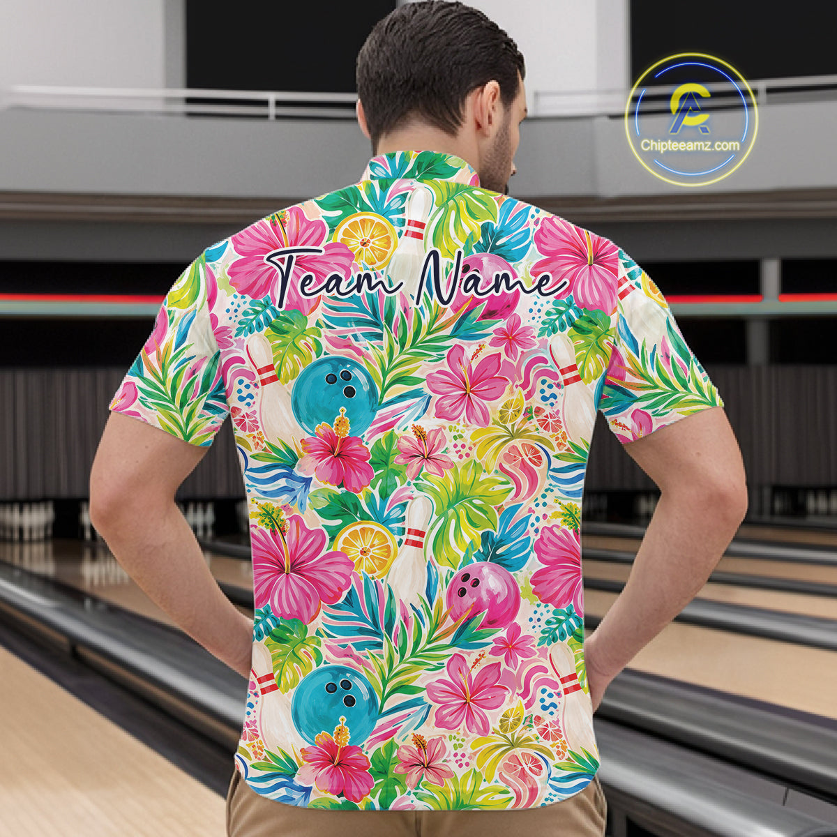 A man viewed from behind wearing a short-sleeve quarter zip shirt with a colorful tropical floral print and bowling-themed graphics, standing in front of bowling lanes. 
