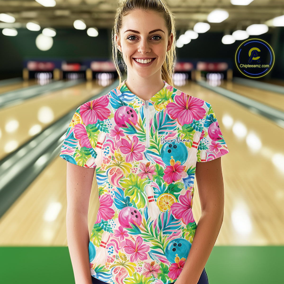 A woman stands in a bowling alley wearing a short-sleeve quarter zip shirt with a bright tropical floral print featuring pink hibiscus flowers, green palm leaves, blue bowling balls