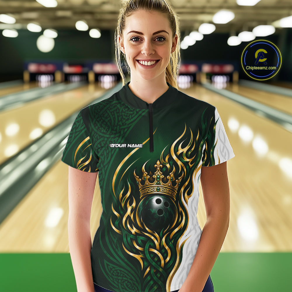 A woman wearing a custom bowling quarter zip shirt jersey stands in a bowling alley. The jersey is dark green with gold flame patterns, a black bowling ball graphic under a gold crown, and white side panels.