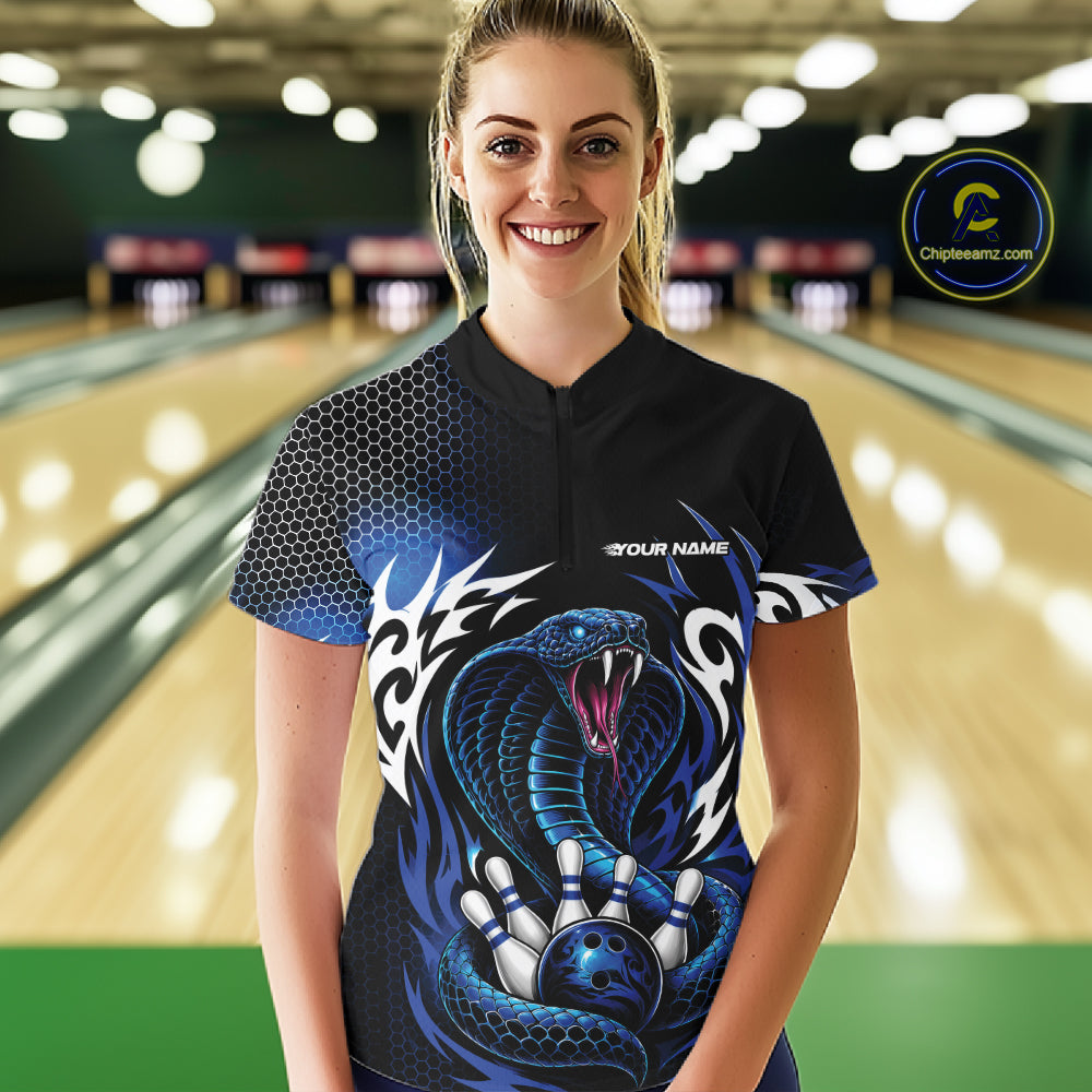 A woman wearing a black bowling quarter zip shirt with a blue and black cobra graphic, standing in a bowling alley. The shirt features a black and blue honeycomb pattern on the sleeves, a blue and black cobra snake graphic on the front