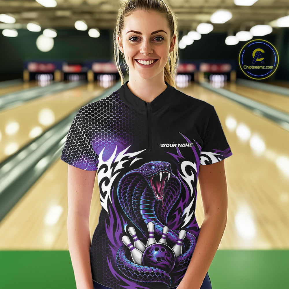 A woman wearing a black bowling quarter zip shirt with a purple and white cobra graphic, standing in a bowling alley 