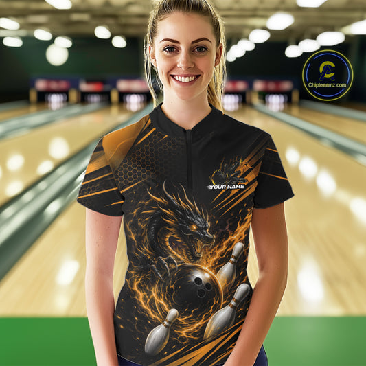 A woman stands in a bowling alley wearing a black short-sleeve bowling shirt with a fiery dragon and bowling ball graphic. The shirt features orange and yellow flame patterns, white bowling pins, and a small white 'YOUR NAME' placeholder on the chest.