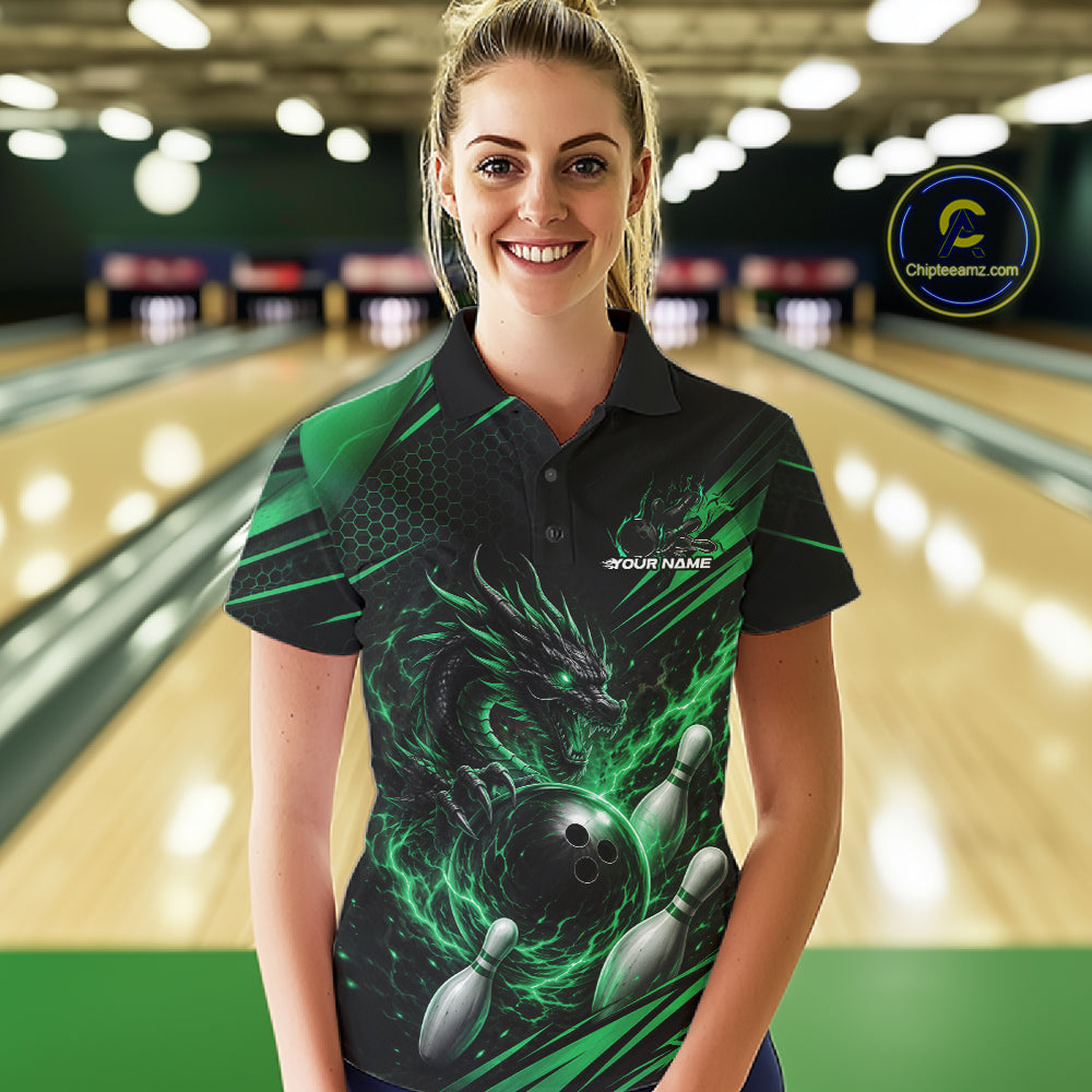 A woman stands in a bowling alley wearing a black short-sleeve polo shirt with a green dragon and bowling ball graphic.