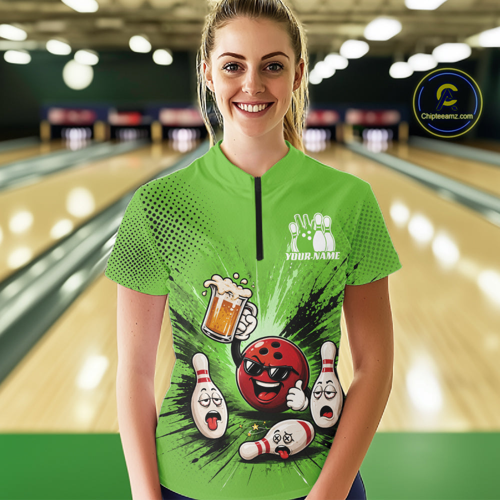 A woman stands in a bowling alley wearing a bright green short-sleeve quarter-zip shirt with a bowling-themed graphic print featuring a red bowling ball, white pins, and a beer mug. 