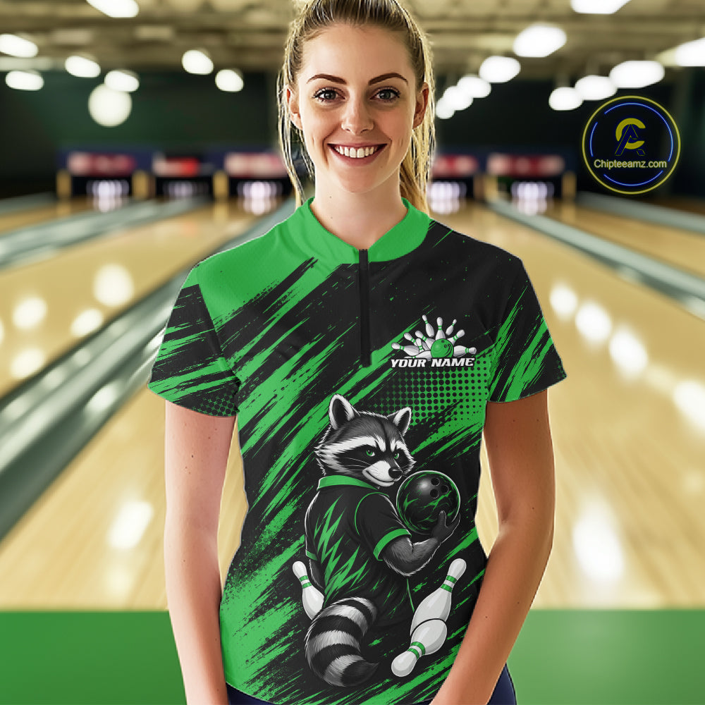 A woman stands in a bowling alley wearing a custom short-sleeve bowling shirt. The shirt features a bold black and green brushstroke pattern with a cartoon raccoon holding a bowling ball on the front.