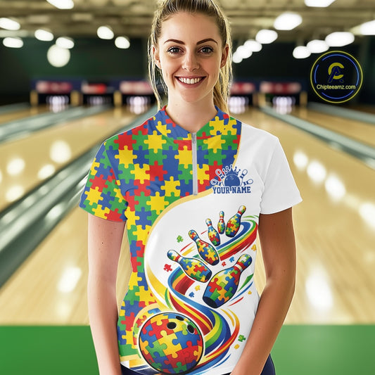 Female model in a bowling lane wearing a white quarter-zip shirt, colorful autism bowling artwork with puzzle-inspired strike visuals.