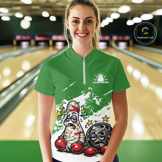 Female model at a bowling lane wearing a green and white quarter-zip shirt, funny boxing-style bowling ball and pin artwork with comedic energy.