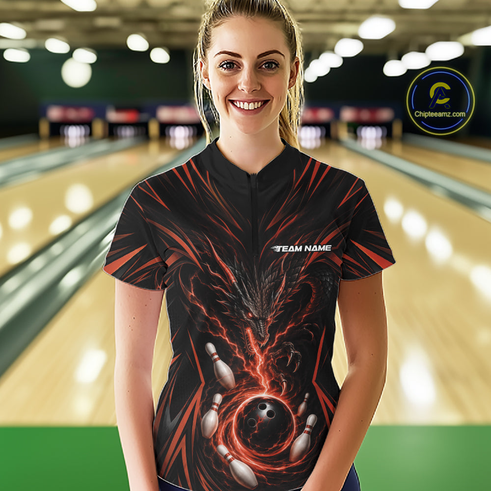 A woman wearing a black bowling-themed short-sleeve quarter zip shirt with orange lightning and bowling pin graphics, standing in a bowling alley. The shirt features a mock collar and a white 'TEAM NAME' placeholder on the chest.