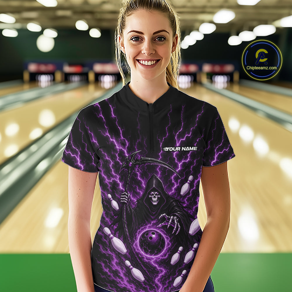 A woman wearing a black bowling-themed quarter zip shirt with a purple grim reaper and bowling ball graphic, standing in a bowling alley with blurred lanes in the background.