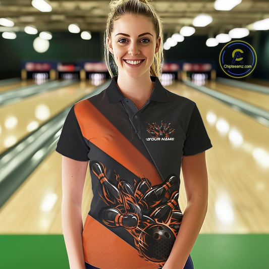Female model front view in a bowling lane wearing a black and orange polo shirt featuring flame bowling ball striking pins in dynamic sports artwork.