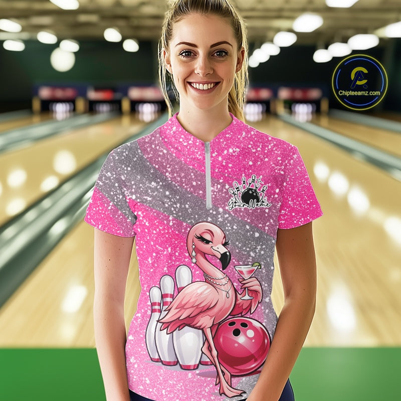 Female model at a bowling lane wearing a pink bowling quarter-zip shirt with an elegant flamingo, cocktail, and bowling ball artwork.