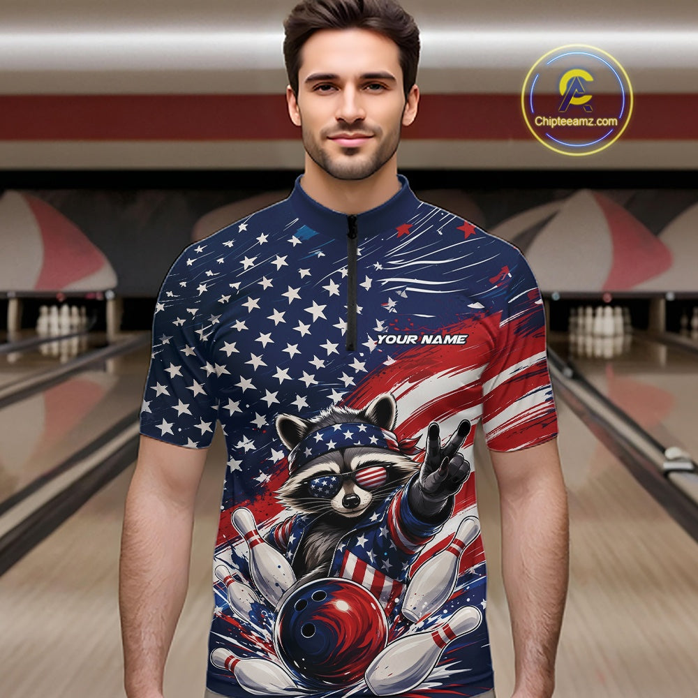 Front view of a male model at a bowling lane wearing a bowling quarter-zip shirt with a patriotic raccoon flashing a peace sign as a red, white, and blue bowling ball crashes through falling pins beneath a flowing American flag.