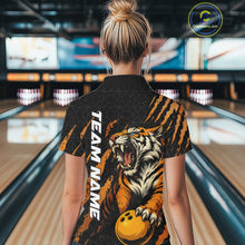 Load image into Gallery viewer, Back view of a female model in a bowling lane wearing a black and yellow bowling polo shirt featuring a roaring tiger holding a yellow bowling ball with orange claw-stripe graphics and a geometric background pattern.