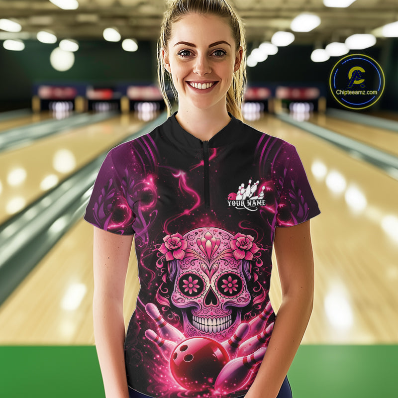 Female model at a bowling lane wearing a black and pink bowling quarter-zip shirt featuring a floral sugar skull above a bowling ball and bowling pins in Day of the Dead style.