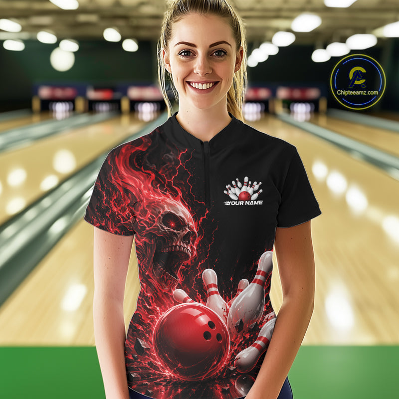 Front view of a female model in a bowling alley wearing a black bowling quarter zip shirt featuring a red fiery skull with lightning striking a bowling ball and pins and a custom name.
