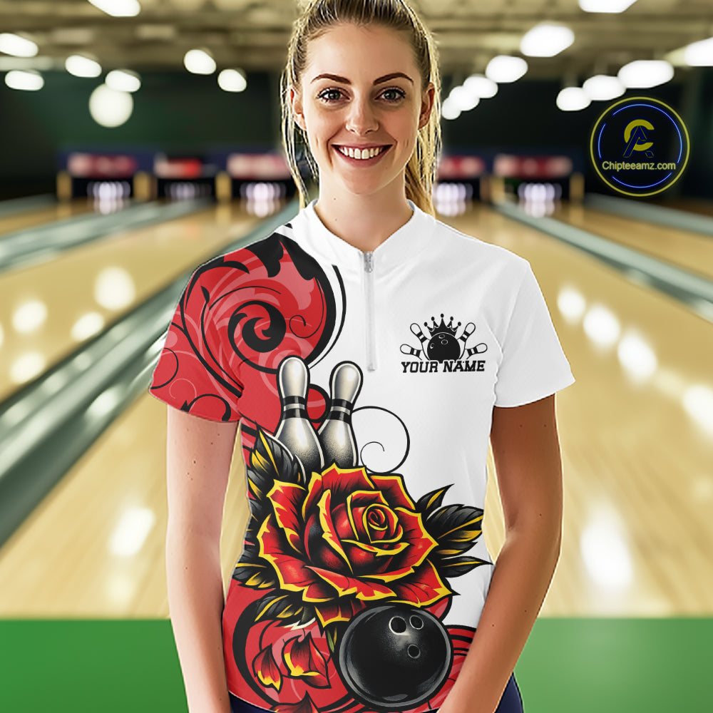 A woman stands in a bowling alley wearing a white short-sleeve bowling quarter zip shirt with a red and black floral design, bowling pins, and a black bowling ball graphic. 