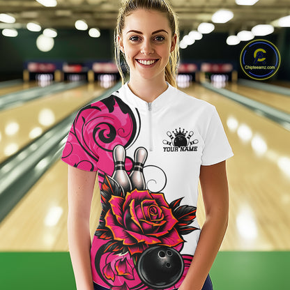 A woman stands in a bowling alley wearing a white short-sleeve bowling quarter zip shirt with a colorful pink and black floral graphic, bowling pins, and a bowling ball design. 
