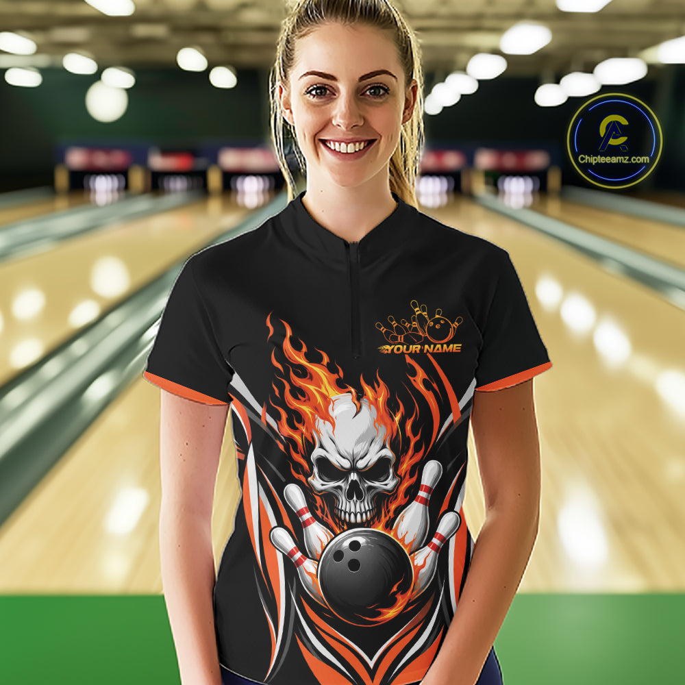 A woman stands in a bowling alley wearing a black short-sleeve bowling quarter zip shirt with a large skull-and-flame graphic and orange trim. The shirt has a mock collar, a short front zipper, and a printed chest design with the text 'YOUR NAME'.
