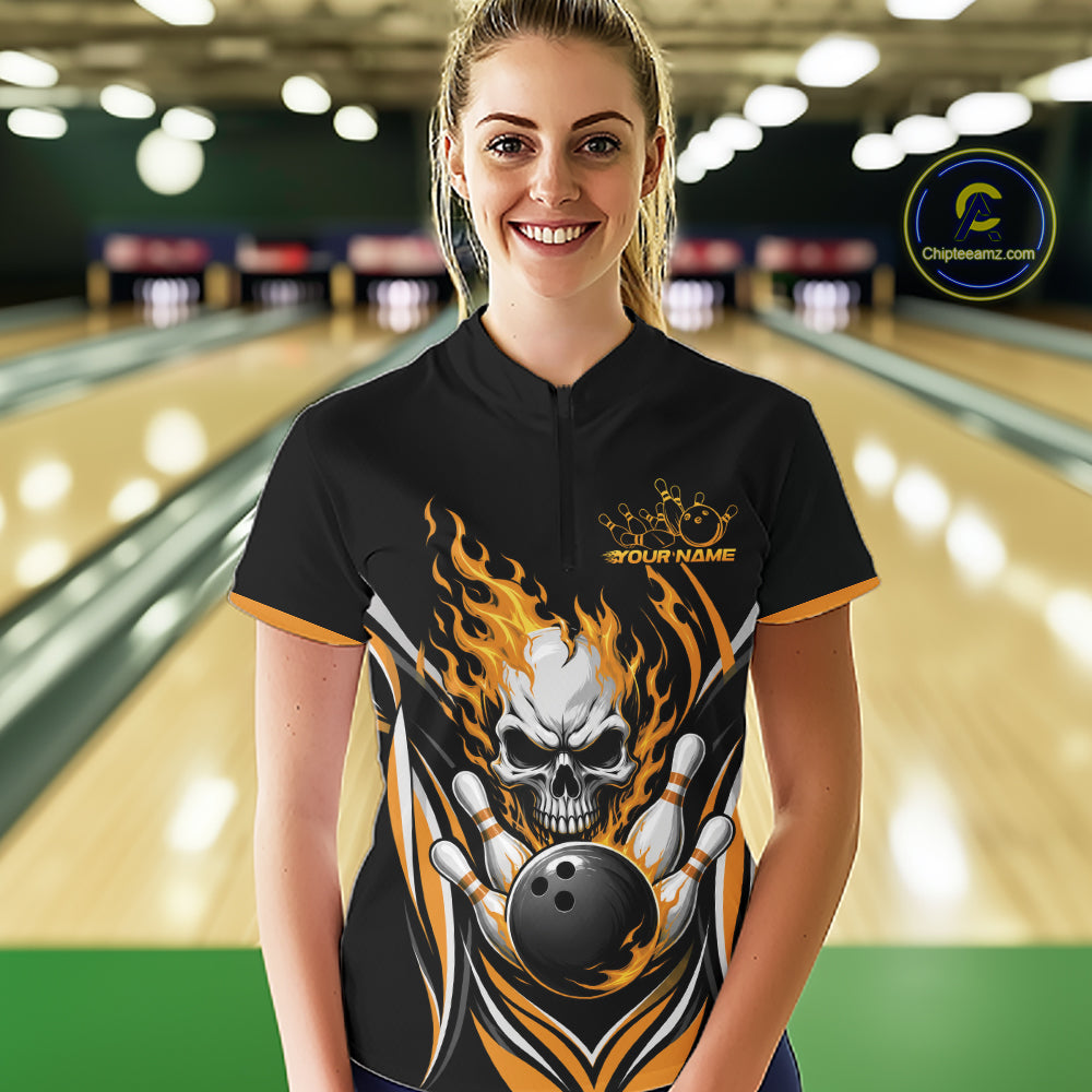 A woman stands in a bowling alley wearing a black short-sleeve bowling quarter zip shirt with a large skull-and-flame graphic and gold trim. 