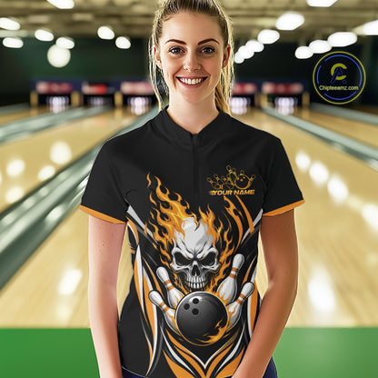 A woman stands in a bowling alley wearing a black short-sleeve bowling quarter zip shirt with a large skull-and-flame graphic and gold trim. 