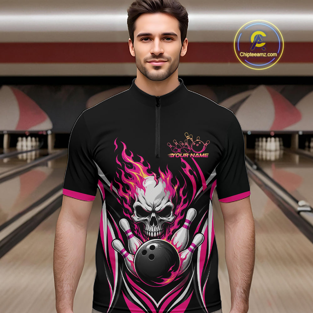 A man wearing a black short-sleeve bowling quarter zip shirt with a large skull and bowling ball graphic in pink, white, and gray, standing in a bowling alley.