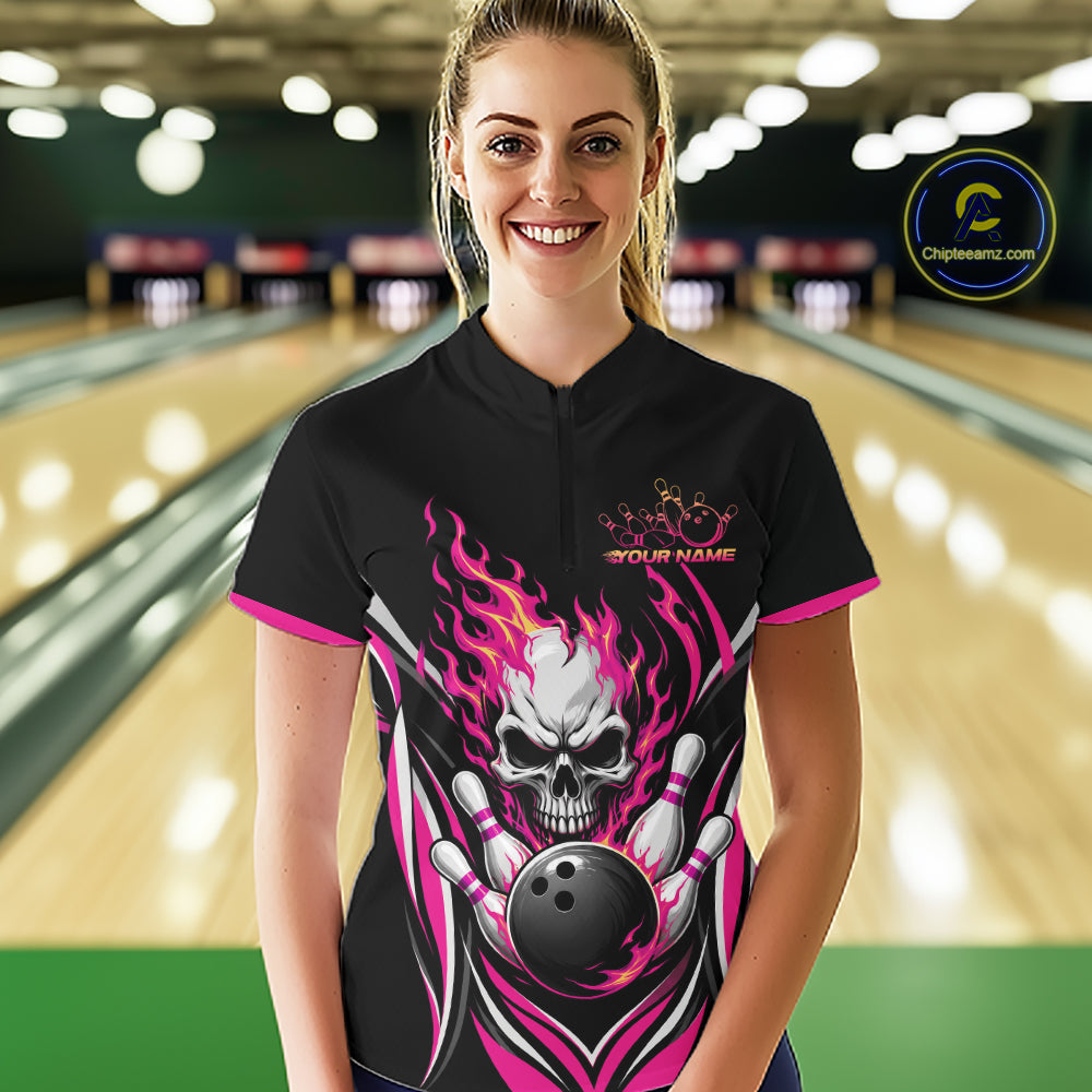 A woman stands in a bowling alley wearing a black short-sleeve bowling quarter zip shirt with a large skull-and-bowling-ball flame graphic and pink trim. 