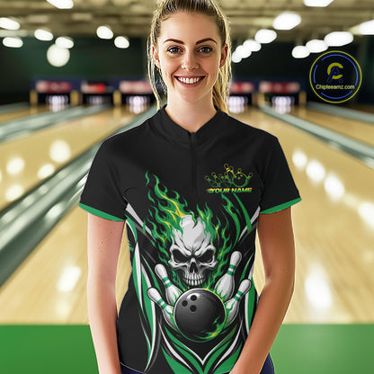 A woman stands in a bowling alley wearing a black short-sleeve bowling quarter zip shirt with a large skull-and-bowling-ball flame graphic on the front and green trim. 