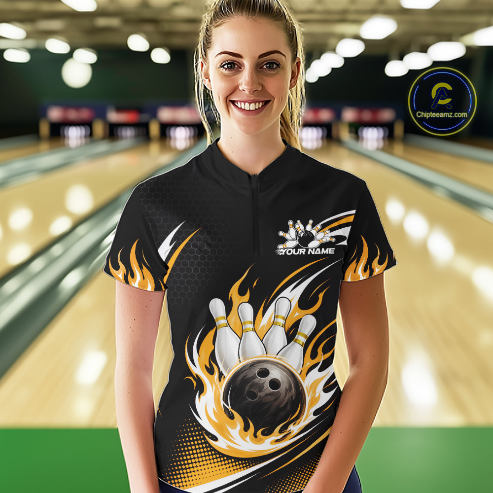 A woman wearing a black bowling-themed short-sleeve quarter zip shirt with a bowling pin and flame graphic, standing in a bowling alley. The shirt features a white and yellow bowling pin design with a bowling ball and flame accents, and a white 'YOUR NAME' placeholder on the chest.