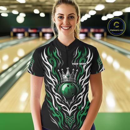 A woman wearing a black short-sleeve bowling quarter zip shirt with a green and white tribal flame design and a crown graphic, standing in a bowling alley. 