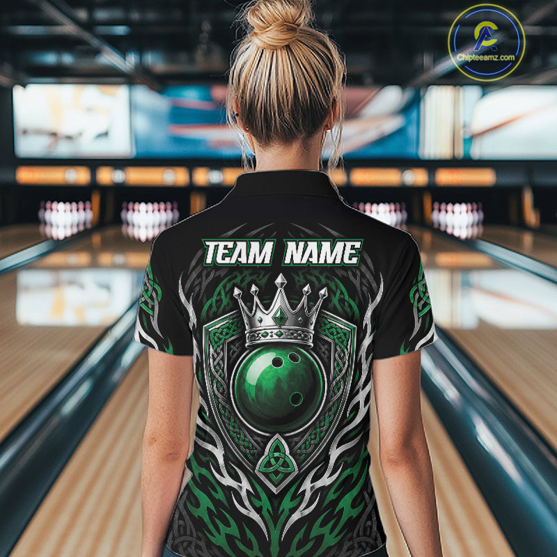 A woman viewed from behind wearing a black bowling short sleeve quarter zip shirt with a large green and crowned bowling ball graphic on the back, standing in a bowling alley.