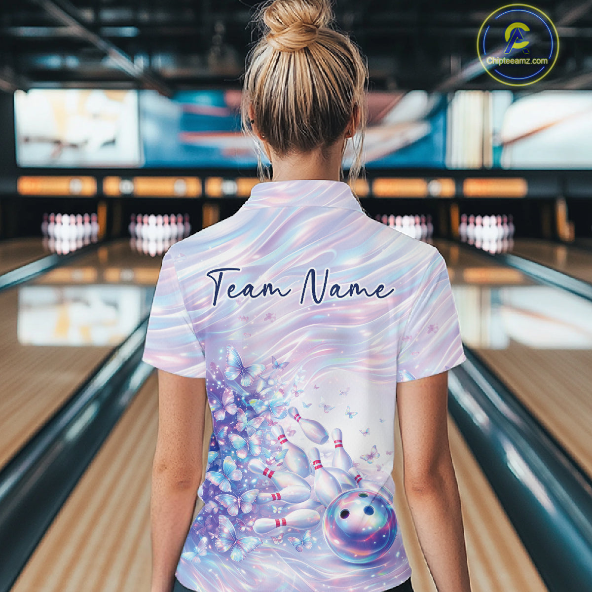 Back view of a female model wearing a bowling quarter zip with pastel purple and pink holographic butterflies and colorful bowling graphics in a bowling alley