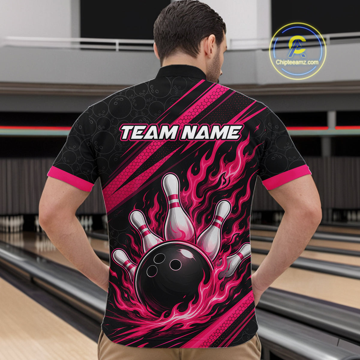 A man viewed from behind wearing a black bowling-themed short-sleeve quarter zip shirt with a vibrant pink and black flame graphic, a bowling ball and pins design on the back, and 'TEAM NAME' printed across the upper back. The background shows a bowling alley with lanes.