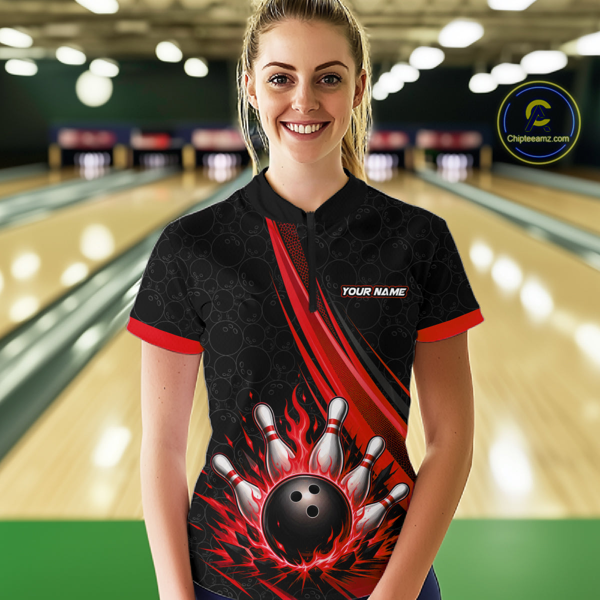 A woman stands in a bowling alley wearing a black short-sleeve quarter zip shirt with red trim and a bold red, black, and white bowling-themed graphic on the front. 
