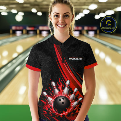 A woman stands in a bowling alley wearing a black short-sleeve quarter zip shirt with red trim and a bold red, black, and white bowling-themed graphic on the front. 
