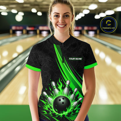 A woman stands in a bowling alley wearing a black short-sleeve quarter zip shirt with bright green flame-like graphics, bowling pins, and a bowling ball design on the front, plus a visible 'YOUR NAME' customization area. 
