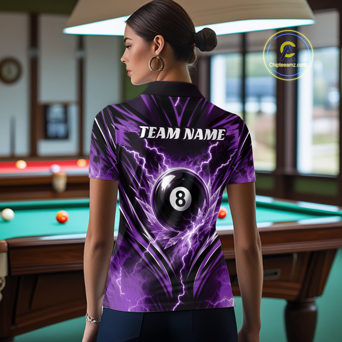 A woman stands in a billiards room wearing a short-sleeve polo shirt from the back. The shirt features a purple and black lightning design with a large 8-ball graphic and the text 'TEAM NAME'.