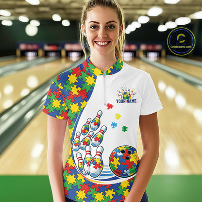 Female model in a bowling lane wearing a white quarter-zip shirt, vibrant autism puzzle bowling artwork with energetic strike-style visuals.
